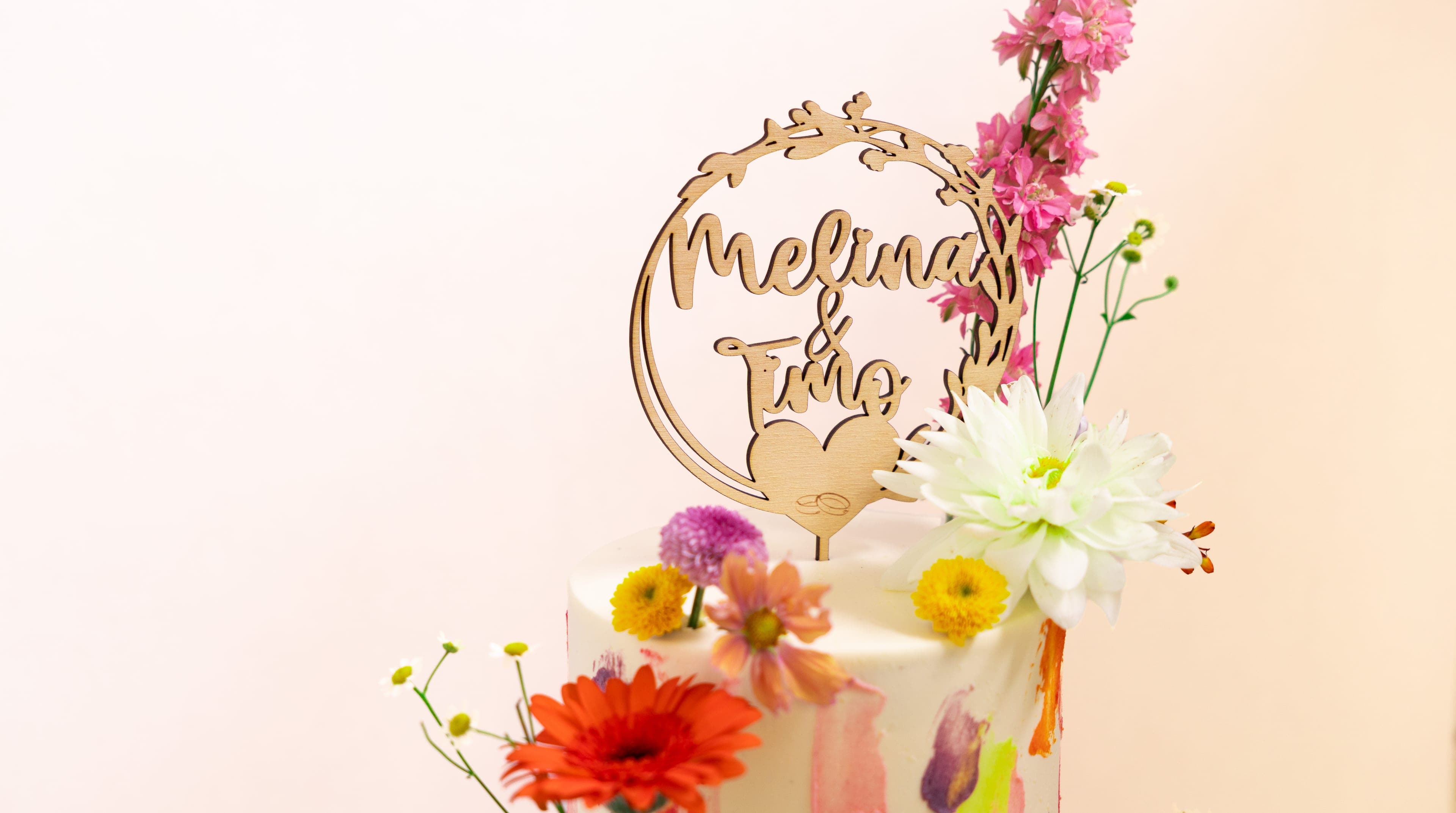 Runder Holz-Topper in Schreibschrift über einer Hochzeitstorte, daneben frische Sommerblüten in Pink, Weiß und Gelb