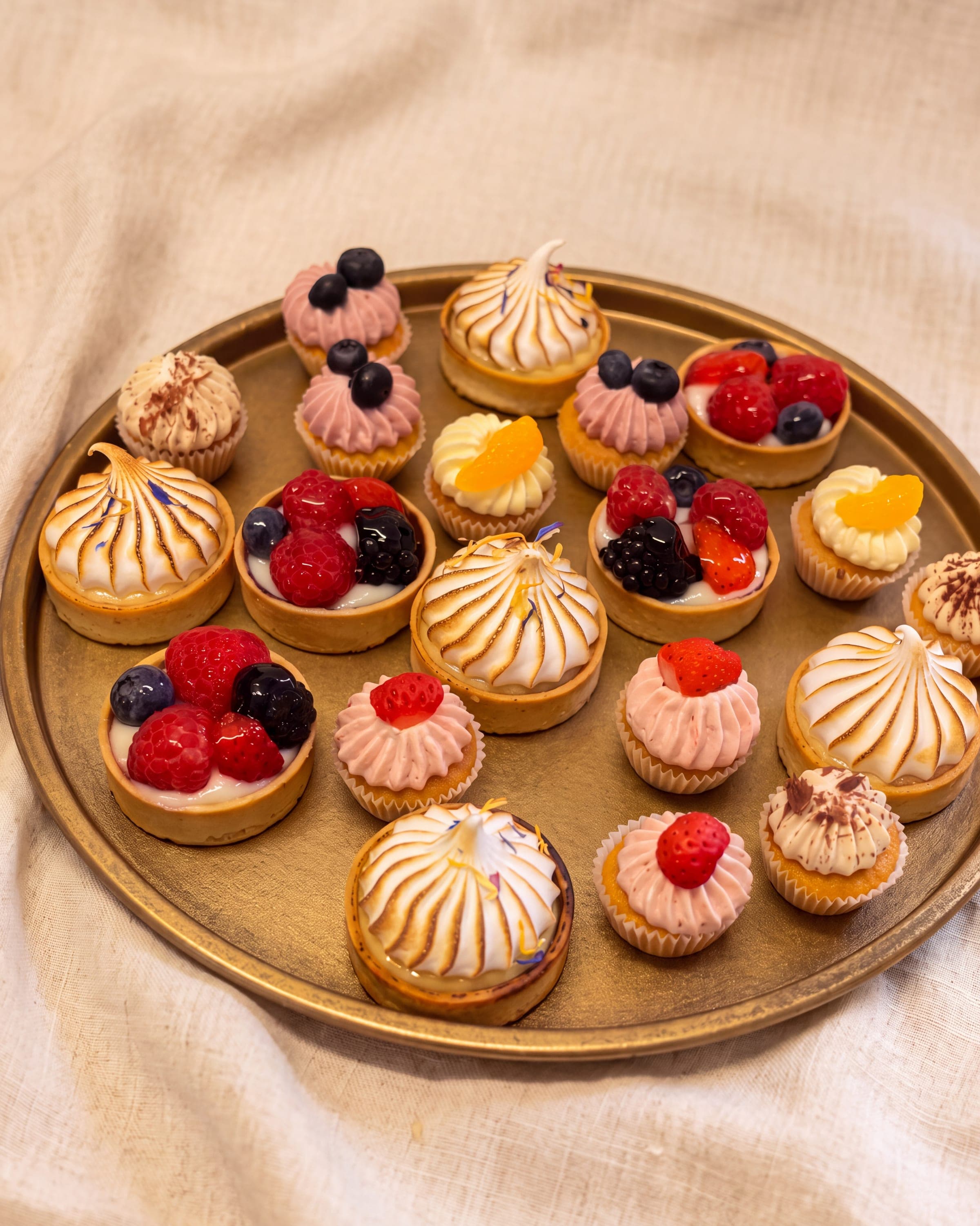 Tartelettes und Törtchen mit Baiserhauben, Beeren und rosa Cremetuffs auf einem goldenen Tablett